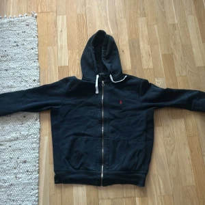 Svart hoodie från Ralph Lauren - Säljer en svart hoodie från Ralph Lauren med dragkedja och huva. Den har en liten röd logotyp på bröstet och är perfekt för en avslappnad stil. Storlek L.