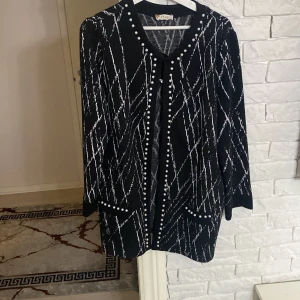 Svart och vit mönstrad kappa - Säljer en elegant svart kappa med vitt mönster och pärldetaljer. Den har långa ärmar och en stilren design som passar perfekt för höst och vinter. Kappan är i bra skick och har en normal passform. Perfekt för att ge din outfit en chic touch!