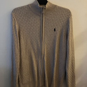 Beige kofta från Ralph Lauren - Snygg beige kofta från Ralph Lauren med dragkedja och ribbad krage. Den har ett diskret rutmönster och den klassiska loggan på bröstet. Perfekt för en stilren look.