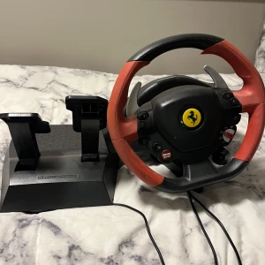 Ratt och pedaler från Thrustmaster - Säljer en Thrustmaster ratt och pedalset med Ferrari-logga. Ratten har en sportig design med svart och röd färg, och pedalerna är svarta. Perfekt för racingentusiaster som vill ha en realistisk körupplevelse hemma. Ansluts med kabel.