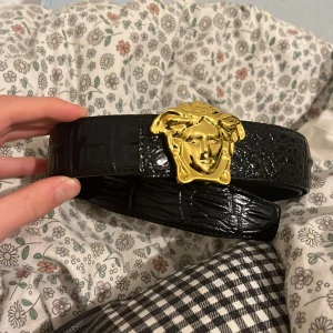 Svart bälte med gulddetalj från Versace - Snyggt svart bälte i skinn från Versace med en iögonfallande guldfärgad spänne i form av ett huvud. Bältet har en texturerad yta som ger en lyxig känsla. Perfekt för att ge en extra touch till din outfit.den är äkta.priset är inte huget i sten skriv ifall ni är intresserade😊