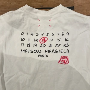 Maison Margiela T-shirt  - Snygg vit t-shirt från Maison Margiela med tryck av siffror och logotyp i svart och rött. T-shirten har en klassisk rund hals och relativt långa ärmar för en T-shirt . Perfekt för en stilren look.