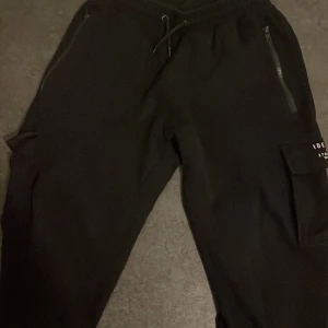 Svarta mjukisbyxor från Woxs - Säljer ett par svarta mjukisbyxor från Woxs i storlek 170. Byxorna har en bekväm passform med resår i midjan och snörning. De har praktiska dragkedjor på fickorna och en cool ficka på sidan med textdetaljer. Perfekta för en avslappnad stil eller träning!