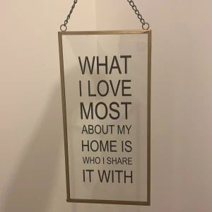 Dekorativ väggskylt med text - Stilren väggskylt med texten 'What I love most about my home is who I share it with'. Skylten har en tunn ram i metall och hängs upp med en kedja. Perfekt för att ge en personlig touch till ditt hem.