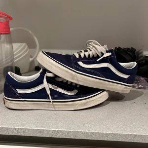 Vans oldskool  - Säljer ett par klassiska blå Vans sneakers med vit sula och snörning. Skorna har den ikoniska vågiga sidodetaljen i vitt och är perfekta för en avslappnad stil. Skorna är i fint skick tycker jag men det ända är att den här röda Vans loggan på hälen är lite sliten på båda skorna men annars ett par riktigt snygga skor