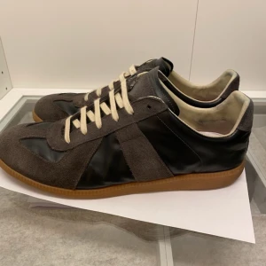 Maison Magiela - Snygga sneakers från Maison Margiela i grå mocka och svart skinn. Skorna har en klassisk design med vita snören och en bekväm gummisula. Perfekta för en stilren look.