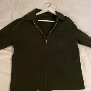 Olivgrön jacka från Riley - Snygg olivgrön jacka från Riley med dragkedja framtill. Jackan har en klassisk krage och två fickor. Perfekt för en stilren look.