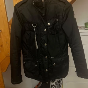 En svart märkes jacka från snoot  - Snygg märkes jacka med både dragkedja och knappar framtill. Jackan har flera fickor och en huva. . Perfekt för kyligare dagar med sin stilrena design och praktiska detaljer.