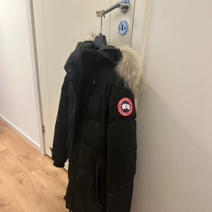  Canada Goose jacka  - Säljer en svart jacka från Canada Goose med pälsdetalj på huvan. Jackan har en klassisk design med lång ärm och dragkedja framtill. Perfekt för kalla vinterdagar. Modellen på jackan heter shelburne. Storlek S