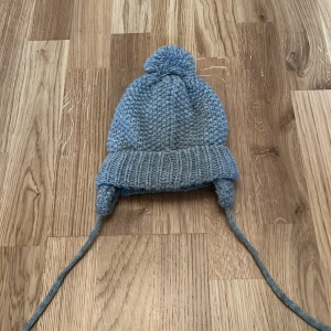 Grå stickad mössa från Crossbow - Mysig grå stickad mössa med tofs och öronlappar från Crossbow. Perfekt för kalla dagar med sitt varma och bekväma material.