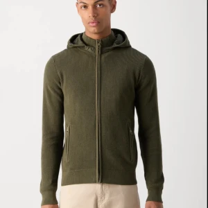 Slutsåld Indicode Cardigan - Snygg och extremt eftertraktad: olivgrön stickad cardigan med dragkedja och huva. Perfekt för kyliga dagar med sin ribbade struktur och praktiska fickor med dragkedja. En stilren och bekväm tröja för alla tillfällen. Slutsåld överallt