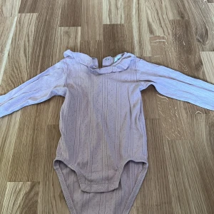 Ljusrosa långärmad body från H&M - Söt ljusrosa långärmad body från H&M med diskret mönster och knappar vid halsen. Perfekt för små barn, tillverkad i mjukt material för bekväm passform.