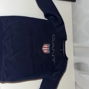 Mörkblå sweatshirt från GANT - Snygg mörkblå sweatshirt från GANT med ett stilrent tryck av märkesloggan och ett emblem på bröstet. Perfekt för en avslappnad och trendig look. 