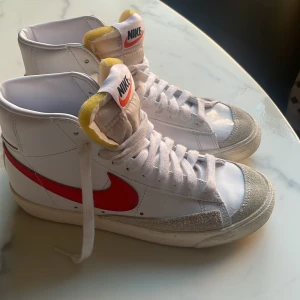 Nike Blazer Mid sneakers - Säljer ett par Nike Blazer Mid sneakers i vitt med röda detaljer. Skorna har en klassisk hög design med snörning och en bekväm passform. De är i bra skick med lite slitage på sulan. Perfekta för både vardag och träning!