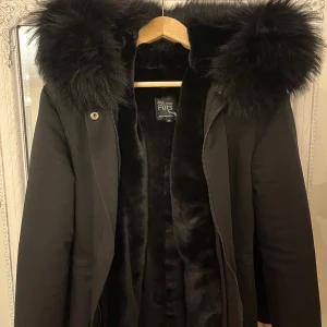 Svart parkajacka med päls - Säljer en snygg svart parkajacka från Wild Furs. Jackan har en stor huva med mjuk päls och är perfekt för kalla vinterdagar. Den har dragkedja och knappar framtill samt en justerbar midja för bästa passform. Insidan är fodrad med mjukt material för extra värme.