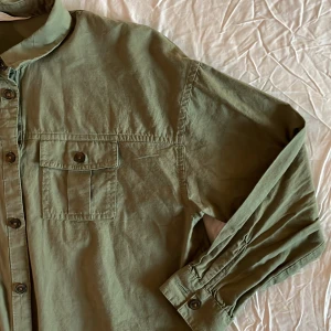 Olivgrön skjorta med knappar - Säljer en snygg olivgrön overshirt i bomull. Den har långa ärmar och två bröstfickor med knappar. Perfekt för lager-på-lager-stil under hösten eller våren. Skjortan har en avslappnad passform och är i mycket bra skick. Perfekt för en casual look!