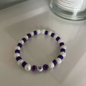 Armband med pärlor och bokstäver - Säljer ett supergulligt armband med vita pärlor och lila detaljer. Armbandet har bokstäverna 'BUU' i mitten, vilket ger det en personlig touch. Perfekt för att piffa upp din outfit eller ge bort som en present!