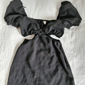 Svart klänning från H&M Divided - Säljer en superfin svart klänning från H&M Divided. Den har puffiga kortärmar och snygga cutouts under bysten, perfekt för både vardag och fest. Klänningen har en smickrande passform med en elastisk midja för extra komfort.