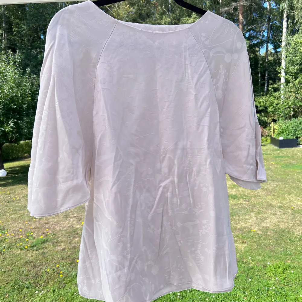 En glansig crémevit blus med vida korta ärmar. Blusen är i storlek 38 och kommer från Anna Glover x H&M. Mycket sparsamt använd! Längd: 57 cm. Puserot.