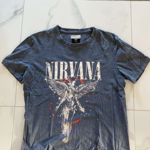 Grå Nirvana t-shirt från Bershka - Säljer en grå Nirvana t-shirt från Bershka. Den har ett coolt tryck med bandets namn och en ängel på framsidan. Perfekt för alla Nirvana-fans! T-shirten är kortärmad och gjord i mjuk bomull, vilket gör den superbekväm att ha på sig. Passar perfekt till vardags eller en avslappnad festkväll.