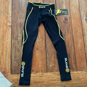 Svarta kompressionstights från Skins - Säljer ett par helt nya svarta kompressionstights från Skins, modell A200. De är designade för träning och har gula detaljer längs sidorna. Perfekta för att ge extra stöd och komfort under intensiva träningspass. Skick 10/10  ( Aldrig använda )