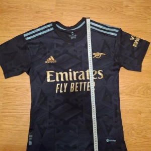 Arsenal 23/24 tröja - Säljer en snygg fotbollströja från Adidas med guldiga detaljer, Arsenal och Adidas märket är broderat. Tröjan har korta ärmar och ett coolt geometriskt mönster. Perfekt för fotbollsfans som vill ha stil både på och utanför planen! Diskuterbart pris!!