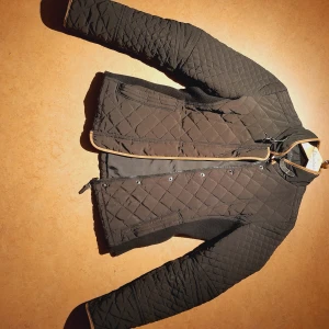 Svart Quiltad Jacka - En svart täckjacka i en lite tight modell, resår i sidorna. Bruna detaljer i skinnimitation. Från Zara i storlek S, skicket är bra! 