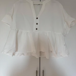 Vit blus med volangdetaljer - Säljer en superfin vit blus med volangdetaljer. Den har korta ärmar och tre knappar framtill som ger en söt touch. Perfekt för vår och sommar, och funkar både till vardags och fest. Passar bra med jeans eller kjol för en chic look!