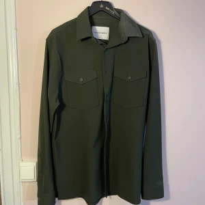 Olivgrön skjorta från Shaping New Tomorrow - Oanvänd! Snygg olivgrön skjorta från Shaping New Tomorrow i storlek M. Skjortan har långa ärmar och två bröstfickor med knappar. Perfekt för både vardag och fest. Materialet känns mjukt och bekvämt, vilket gör den idealisk för alla säsonger. Är mörkare än vad som syns på bilderna. Sista bilden är mest lik i tonen. Fast mörkare
