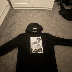 Svart hoodie från Boss med tryck - Säljer en hoodie från Boss som gjort en collab med Muhammed Ali. Är en limiterad vara, kvitto och tags finns. Väldigt sparsamt har mest legat i garderoben Hoodien har en normal passform och är storlek M. Ny pris är 1100:- 
