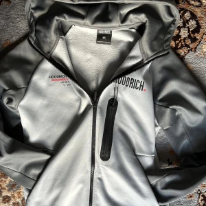 Grå hoodie från Hoodrich - Säljer en snygg grå hoodie från Hoodrich i storlek XS. Den har en dragkedja framtill och en stor huva. På bröstet och ryggen finns tryck med texten 'Hoodrich Lifestyle Worldwide Organisation'. Perfekt för en casual look eller när du vill hålla det bekvämt men stiligt.