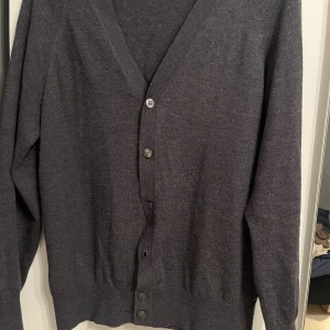 Grå kofta från Tiger of Sweden - Säljer en elegant grå kofta från Tiger of Sweden, tillverkad av fin italiensk garn. Koftan har en klassisk design med knappar framtill och långa ärmar. Perfekt för både vardag och festliga tillfällen. Passar bra till höst och vinter. Storlek XL i barnstorlek men sitter som en vanlig S. Hör av er vid ytterligare frågor!