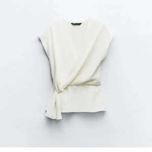 Vit omlottopp från Zara - Snygg vit omlottopp från Zara. Toppen har en knytning i sidan och är ärmlös, vilket gör den perfekt för både vardag och fest. Passar bra till både jeans och kjol!