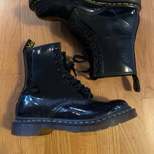 Svarta lackade Dr. Marten - Säljer ett par svarta, glansiga Dr. Marten boots i lackat material. De har en klassisk snörning och en robust sula med gul söm. Perfekta för höst och vinter, och ger en cool touch till vilken outfit som helst. Använda men i mycket bra skick!