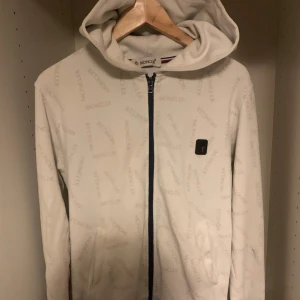  Moncler kofta  - Säljer en snygg vit hoodie från Moncler i mycket bra skick. Den har ett diskret mönster med Moncler-loggan över hela tyget och en dragkedja framtill. Perfekt för höst och vår! 🧥