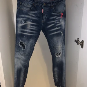 Blå jeans från Dsquared2 - Säljer ett par snygga blå jeans från Dsquared2 med coola slitningar och distressed detaljer. De har en normal passform och är perfekta för en avslappnad stil. Märkesloggan syns tydligt på den röda etiketten vid fickan. Perfekta för både vardag och fest!