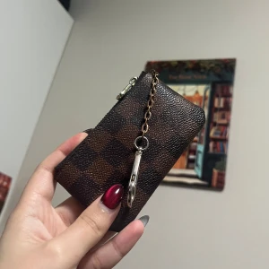 Brun rutig plånbok med kedja - Säljer en snygg brun rutig plånbok med dragkedja och kedjedetalj. Perfekt för att hålla dina kort och småsaker säkra. Den har en praktisk karbinhake för att fästa i väskan eller nyckelringen dock har den låst sig men du kanske vet hur man öppnar den. Perfekt accessoar för  vardagen ! Söks till en billig peng med tanke på att färgen på kedjan ändrats ✨☺️