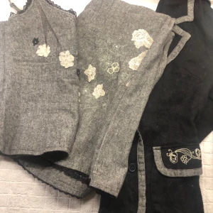 3 delat set om kjol, linne och kavaj - Säljer en elegant svart kavaj med gråa broderade detaljer och knappar framtill. Kavajen har en snygg passform och är perfekt för höst och vår. Den har långa ärmar och dekorativa fickor med broderi. Perfekt för både vardag och fest! Medföljer gör även ett linne och en kjol.