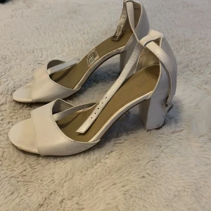 Vita klackskor med öppen tå - Säljer ett par eleganta vita klackskor med öppen tå och justerbart spänne runt ankeln. Perfekta för sommarens fester eller en stilren look till vardags. De har en stabil blockklack som ger bra stöd och komfort hela dagen.