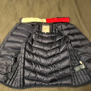 Mörkblå dunväst från Tommy Hilfiger - Säljer en snygg mörkblå dunväst från Tommy Hilfiger. Den har en cool färgblockdesign med röd och vit insida på kragen. Västen är quiltad och har dragkedja framtill samt två praktiska fickor med dragkedjor. Perfekt för höst och vår när du vill hålla dig varm men ändå se stilren ut. ❗️Kom med prisförslag❗️