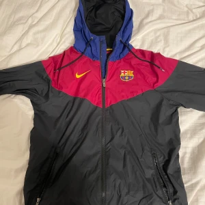 FC Barcelona vindjacka  - Säljer en snygg FC Barcelona vindjacka från Nike. Den har en cool färgkombination av svart, röd och blå med klubbens emblem på bröstet. Jackan har en dragkedja framtill och en huva för extra skydd. Perfekt för fotbollsfans! 🖤❤️💙