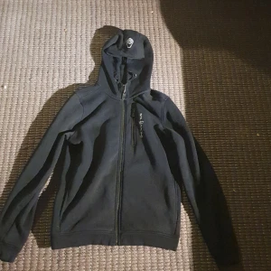 Svart hoodie från Sail Racing (äkta) - Säljer en svart hoodie från Sail Racing i nytt skick. Den har en dragkedja framtill och en praktisk huva. Perfekt för höst och vår, och funkar lika bra till vardags som till träning. Skön och stilren design som passar till det mesta. 🖤