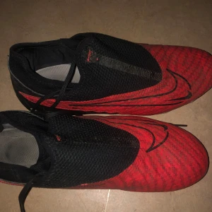 Röda och svarta fotbollsskor från Nike - Säljer ett par coola röda och svarta fotbollsskor från Nike. De har en snygg design med svarta detaljer och är perfekta för fotbollsträning eller match. Skorna har dobbar för bra grepp på planen och snörning för en säker passform.