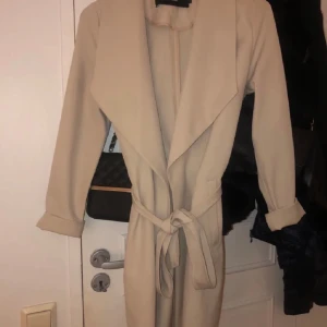 Beige kappa från Vero Moda - Säljer en stilren beige kappa från Vero Moda i storlek S. Den har ett knytbälte i midjan och långa ärmar. Perfekt för höst och vår när du vill ha en elegant look. Kappan är i mycket bra skick och redo för nya äventyr!