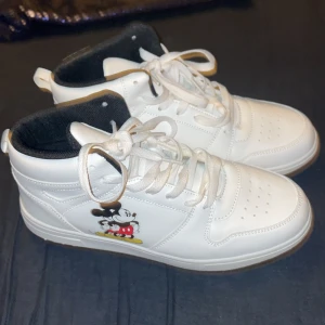 Vita sneakers med Musse Pigg på - Säljer ett par vita sneakers från Disney i storlek 39. Skorna har ett coolt Musse Pigg-motiv på sidan och är i syntetmaterial. De har snörning och en bekväm passform, perfekt för vardagsbruk. Skorna är nya och aldrig använt