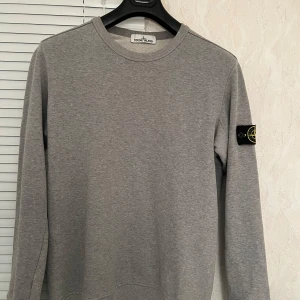 Grå tröja från Stone Island - Säljer en snygg grå tröja från Stone Island i bomull. Den har en klassisk rund hals och deras ikoniska märke på ärmen. Perfekt för både vardag och lite mer avslappnade tillfällen. Tröjan är i mycket bra skick och redo för en ny ägare!