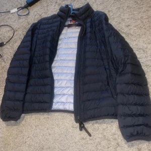 Svart dunjacka från parajumper - Säljer en svart dunjacka från parajumperi bra skick. Den har en dragkedja framtill och är quiltad för extra värme. Perfekt för kyliga höst- och vinterdagar. Jackan har långa ärmar och en snygg logga på ärmen. Insidan är ljusgrå vilket ger en cool kontrast.