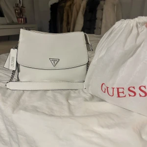 Vit axelväska från Guess - Säljer en snygg vit axelväska från Guess i mycket bra skick. Väskan har en stilren design med en kedjedetalj på axelremmen och Guess-logga på framsidan. Äkta Guess väska! Vit med svarta och silvriga detaljer. Använd Max 3 ggr, man får med dustmagen såklart. Säljer för 699kr, kom gärna med bud!! 
