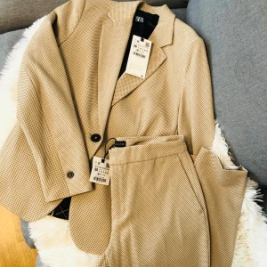 Beige kavaj och byxor från Zara - Säljer en snygg beige kavaj och matchande byxor från Zara i storlek 36. Setet har ett diskret rutigt mönster och är perfekt för både kontoret och festliga tillfällen. Kavajen har en klassisk passform med knappar framtill och byxorna har en bekväm midja. Helt nytt med taggar kvar!