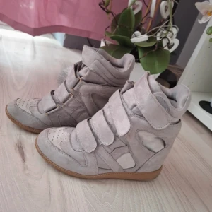 Grå sneakers med kilklack - Säljer ett par grå sneakers med kilklack i mocka. De har en cool design med flera kardborreband för en säker passform. Perfekta för en avslappnad men stilren look. Sulan är i en kontrasterande brun färg som ger en snygg touch.